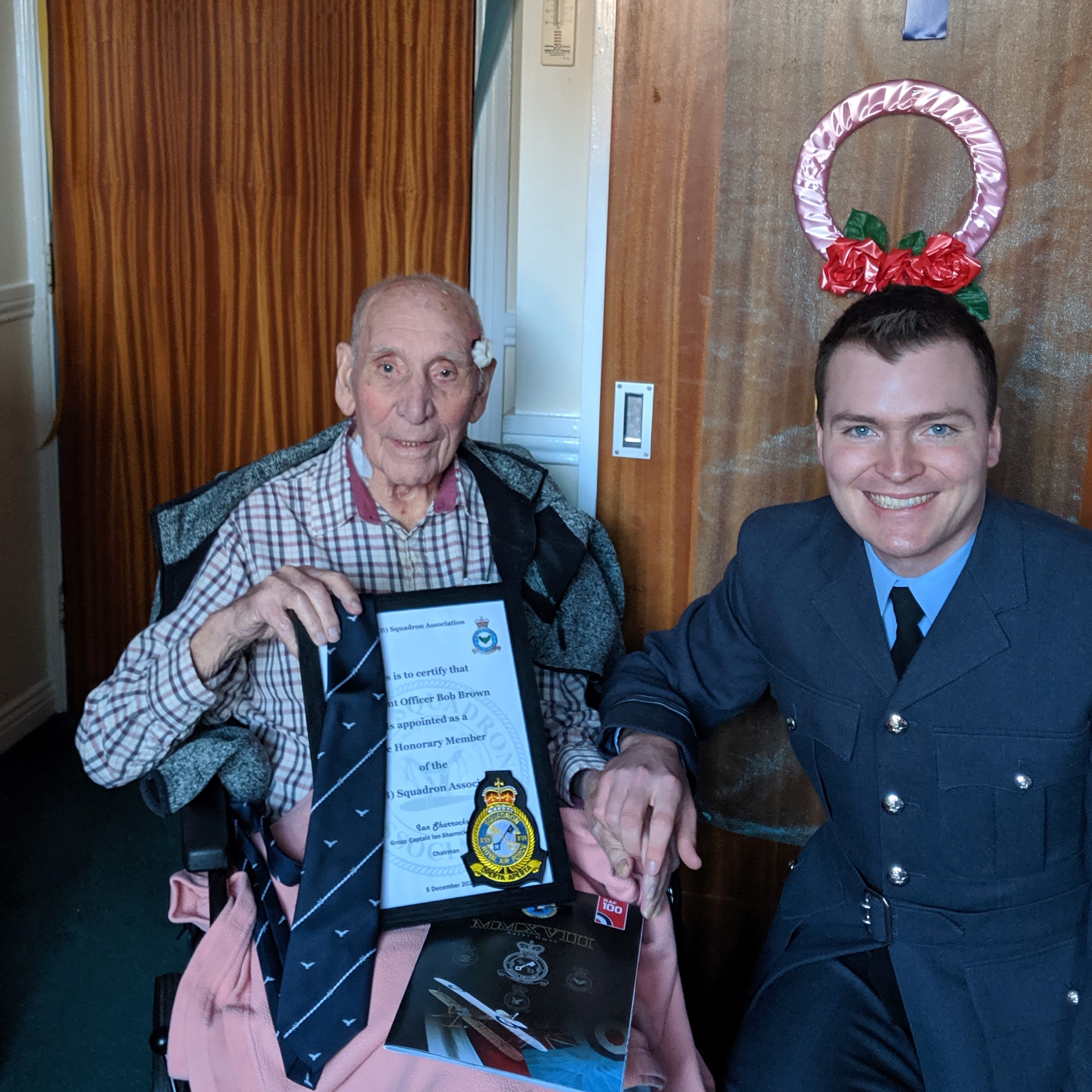 Image shows RAF veteran holding a certificate and a tie while sitting next to a RAF aviator.